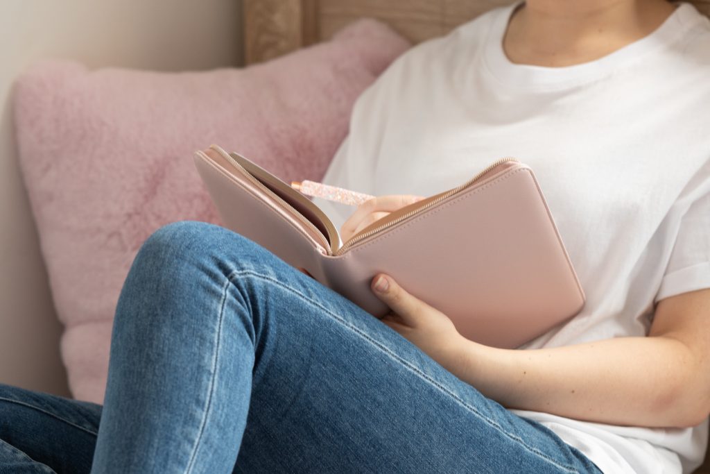 middle view of someone in a white cotton top writing in a pink leather notepad relaxed on a couch.