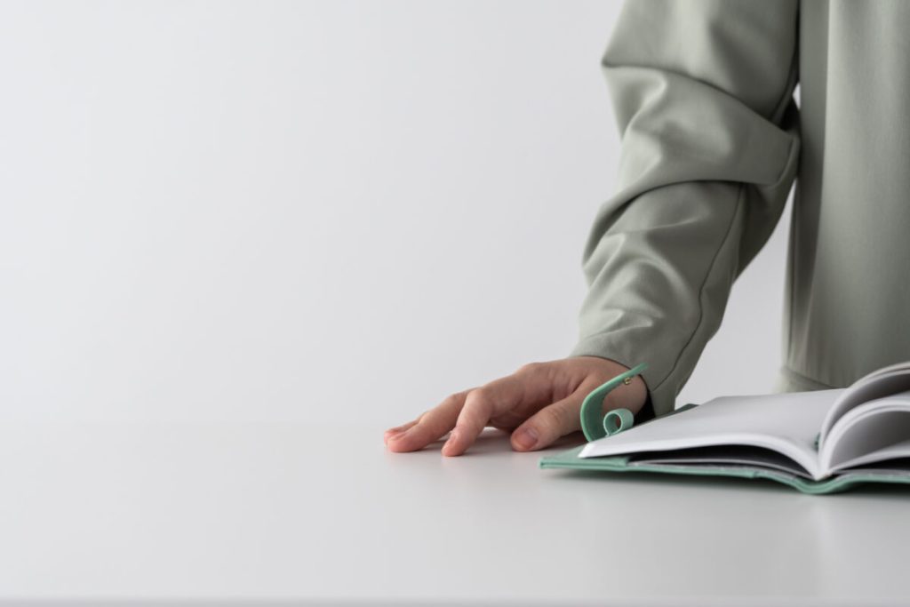 White desk and background, someones arm in a sage long sleeve shirt with their hand next to a sage bound notepad.