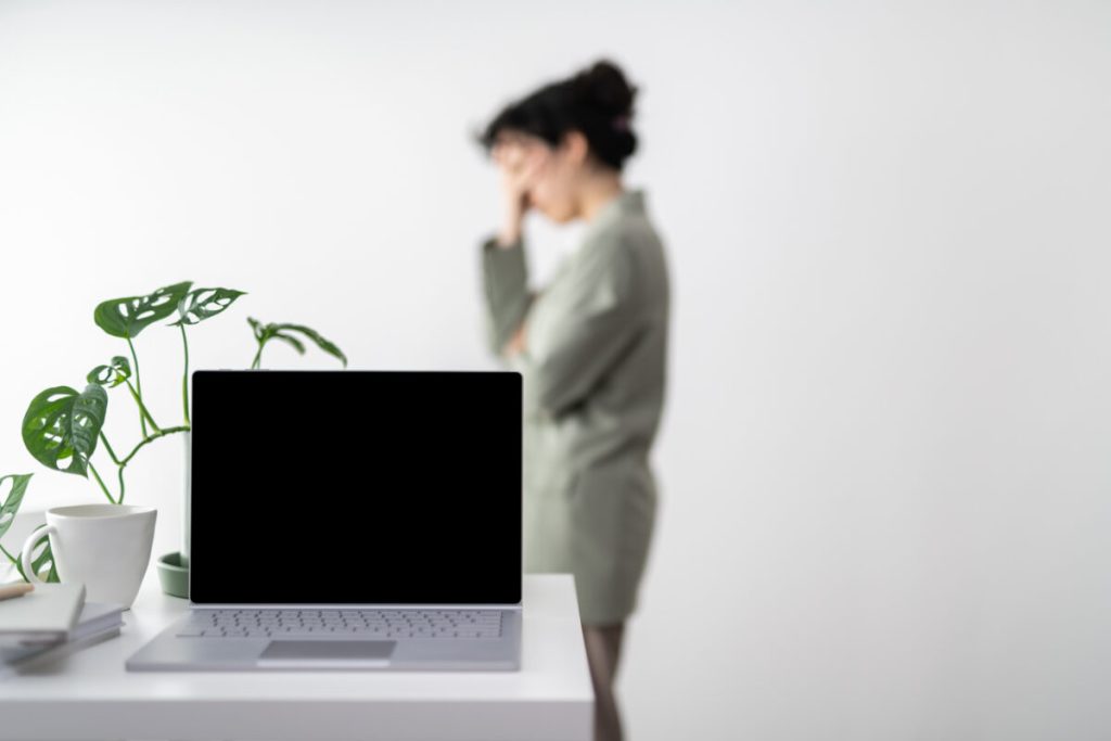 Laptop with black screen on a white desk in the foreground with a swiss cheese vine behind it. A person in a sage shirt blurred in the background looking worried holding their face in their hands.