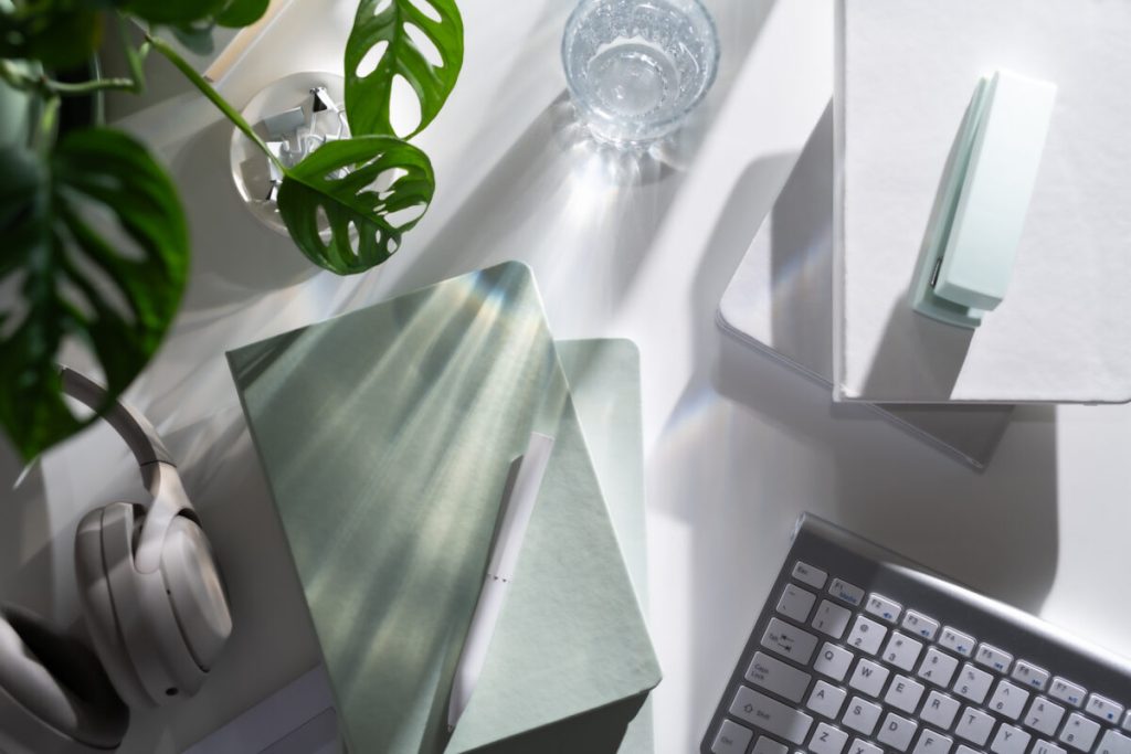 White desk flatlay with a green swiss cheese vine, green notepad and pen, corner of a keyboard and headphones.